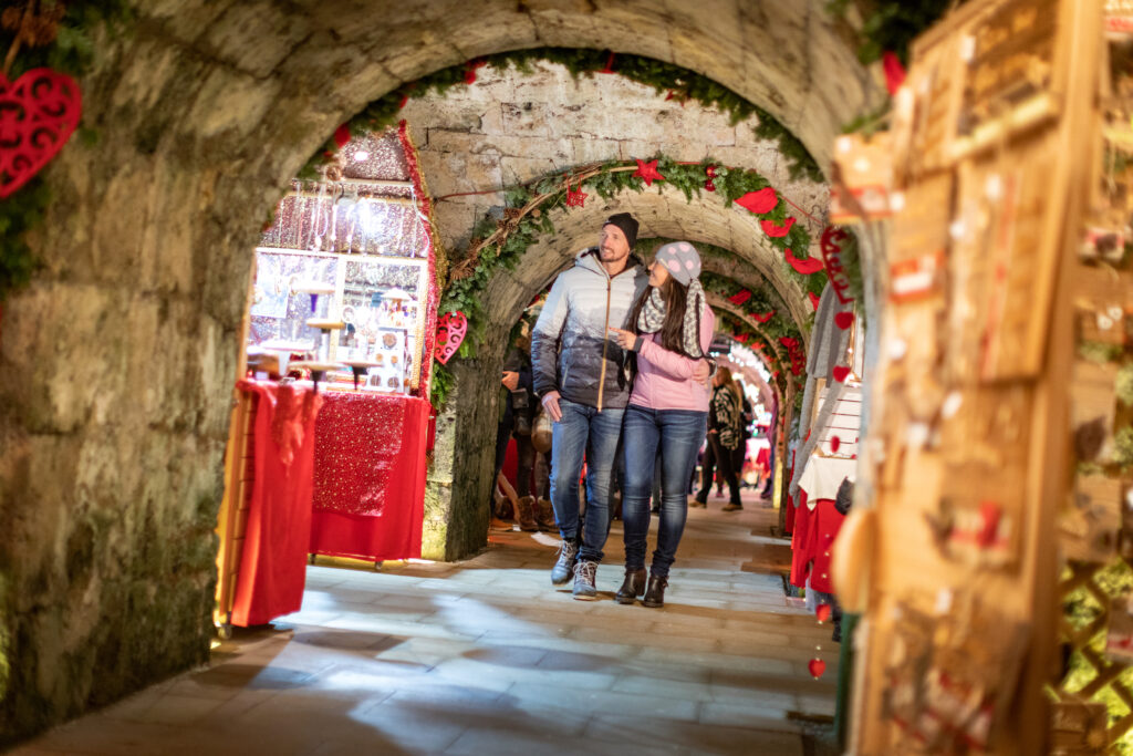 Una coppia cammina attraverso una galleria decorata con ornamenti natalizi e bancarelle al mercatino di Natale sulla Fortezza Kufstein.