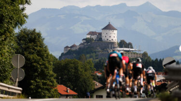 Radfahrer fahren während eines Radrennens bergauf, im Hintergrund ist die Festung Kufstein umgeben von üppigen Bäumen und hügeliger Landschaft.