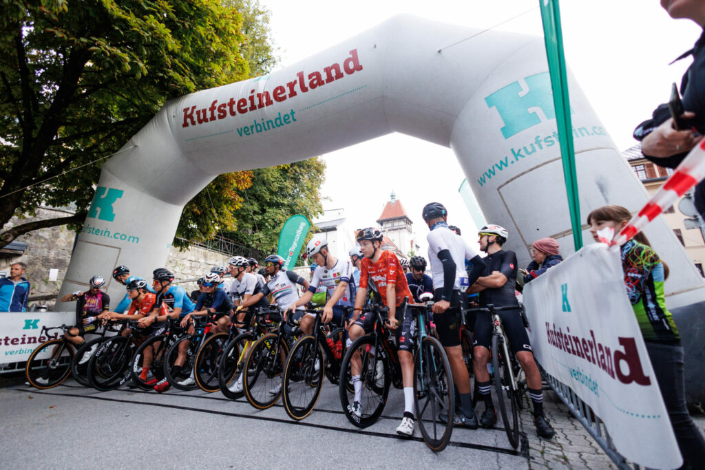 Radfahrer stehen an der Startlinie eines Radrennens in Kufstein unter einem großen aufblasbaren Tor.