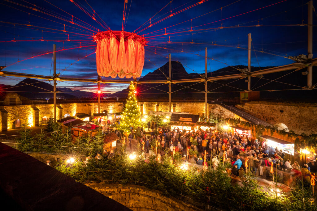 Vista notturna di un mercatino di Natale all'interno di la Fortezza Kufstein, con una grande albero di Natale illuminato e bancarelle circondate da visitatori.