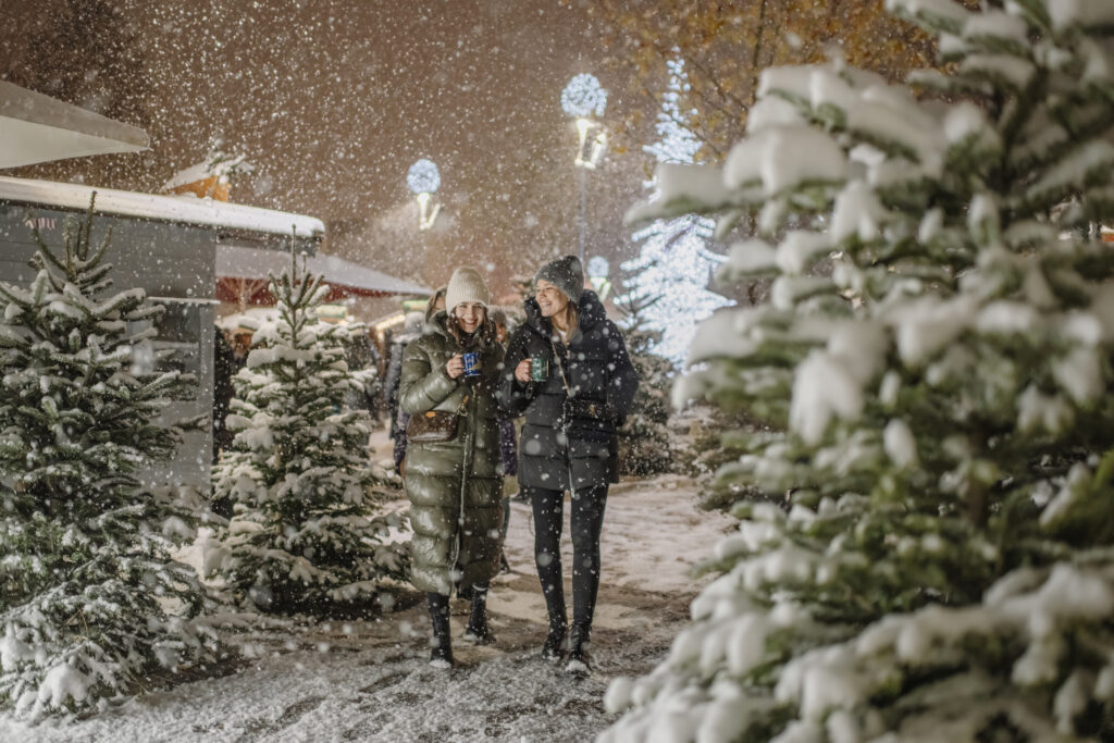 Due amiche che camminano nel mercatino di Natale di Kufstein durante una nevicata, circondate da alberi di Natale illuminati.