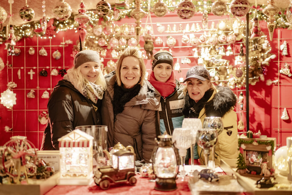 Quattro venditori sorridenti posano davanti alla loro bancarella decorata con ornamenti natalizi al mercatino di Natale di Kufstein.