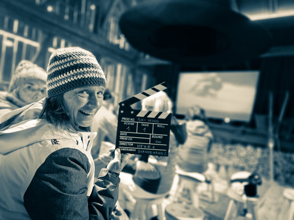 Altes Bauernhaus in dem Hocker in Kinobestuhlung aufgestellt sind, Leinwand mit Nostalgiefilm läuft, im Vordergrund Zuschauer mit Tüte Popcorn