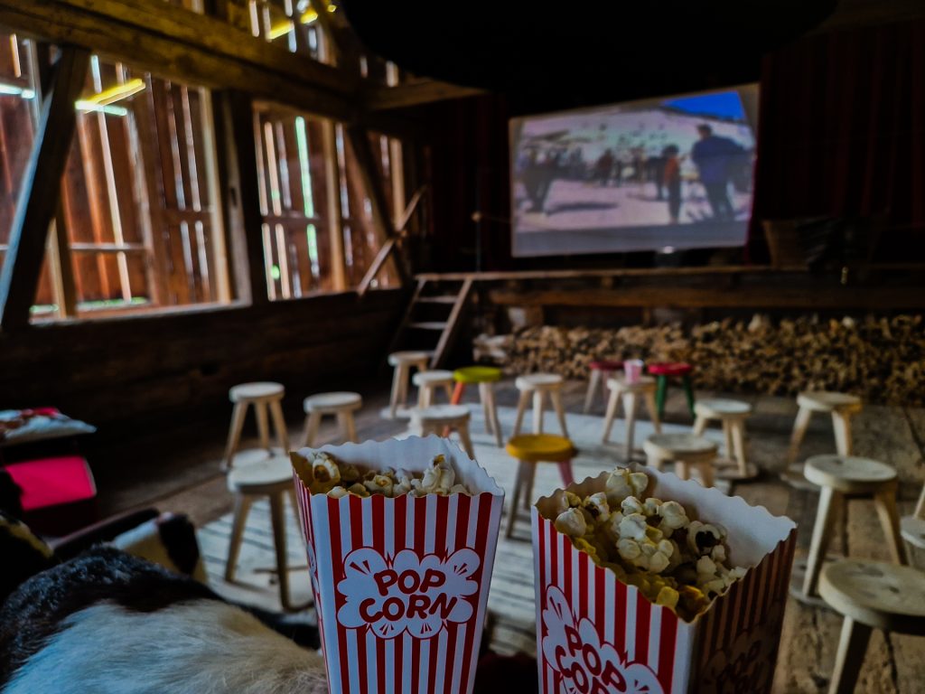 Altes Bauernhaus in dem Hocker in Kinobestuhlung aufgestellt sind, Leinwand mit Nostalgiefilm läuft, im Vordergrund Tüten Popcorn