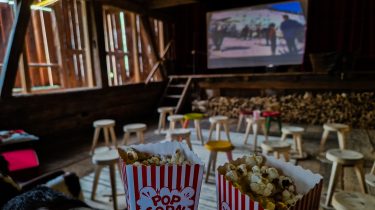 Altes Bauernhaus in dem Hocker in Kinobestuhlung aufgestellt sind, Leinwand mit Nostalgiefilm läuft, im Vordergrund Tüten Popcorn
