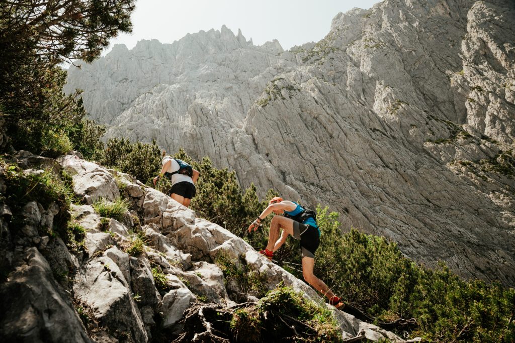 Due trail runner salgono un ripido sentiero roccioso alpino con alte pareti montuose sullo sfondo.