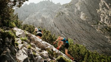 Due trail runner salgono un ripido sentiero roccioso alpino con alte pareti montuose sullo sfondo.