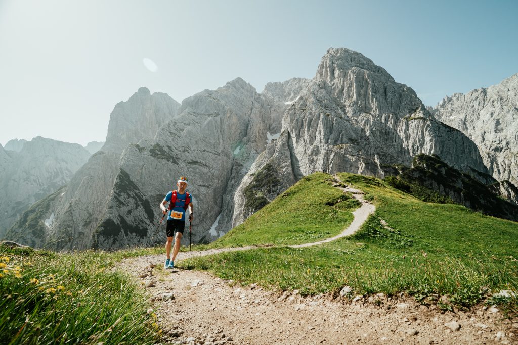 Trail runner con bastoncini su sentiero alpino con spettacolari cime rocciose sullo sfondo.