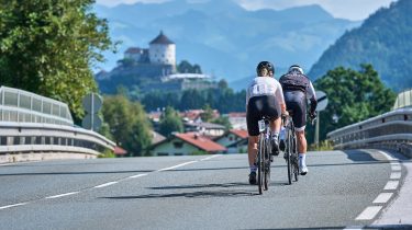 Zwei Rennradfahrer fahren nebeneinander auf einer Straße in Richtung Kufstein, die Festung Kufstein im Hintergrund.