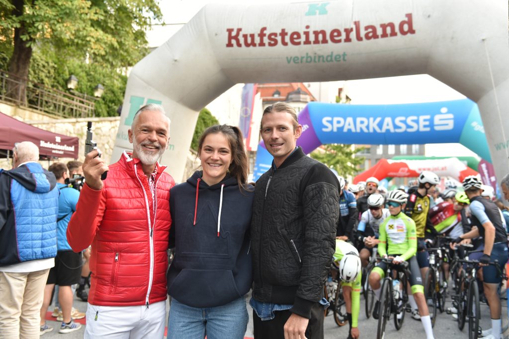Georg Hörhager, Lina Schneeweiß, Thomas Krimbacher posieren vor dem Startbogen eines Radmarathons, während sich dahinter zahlreiche Teilnehmende mit Rennrädern sammeln.