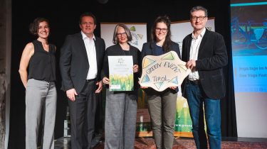 Green Event Tirol - Yoga-Festvial des TVB Kufsteinerland ausgezeichnet. Fünf Personen stehen auf einer Bühne und präsentieren eine Urkunde sowie ein „Green Event Tirol“-Schild bei einer Auszeichnungsveranstaltung.