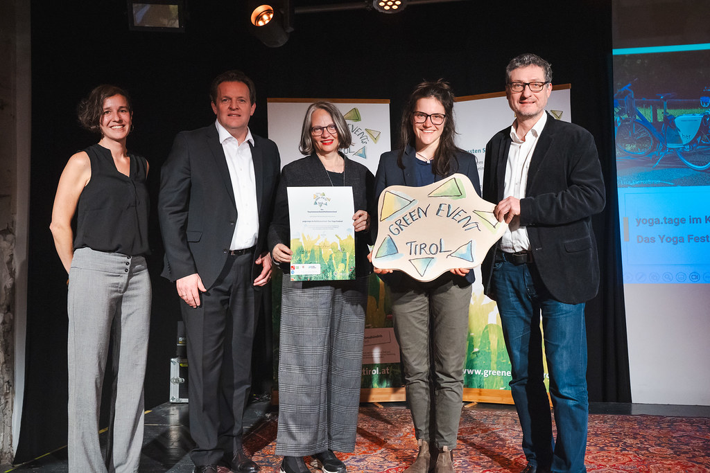 Green Event Tirol - Yoga-Festvial des TVB Kufsteinerland ausgezeichnet. Fünf Personen stehen auf einer Bühne und präsentieren eine Urkunde sowie ein „Green Event Tirol“-Schild bei einer Auszeichnungsveranstaltung.