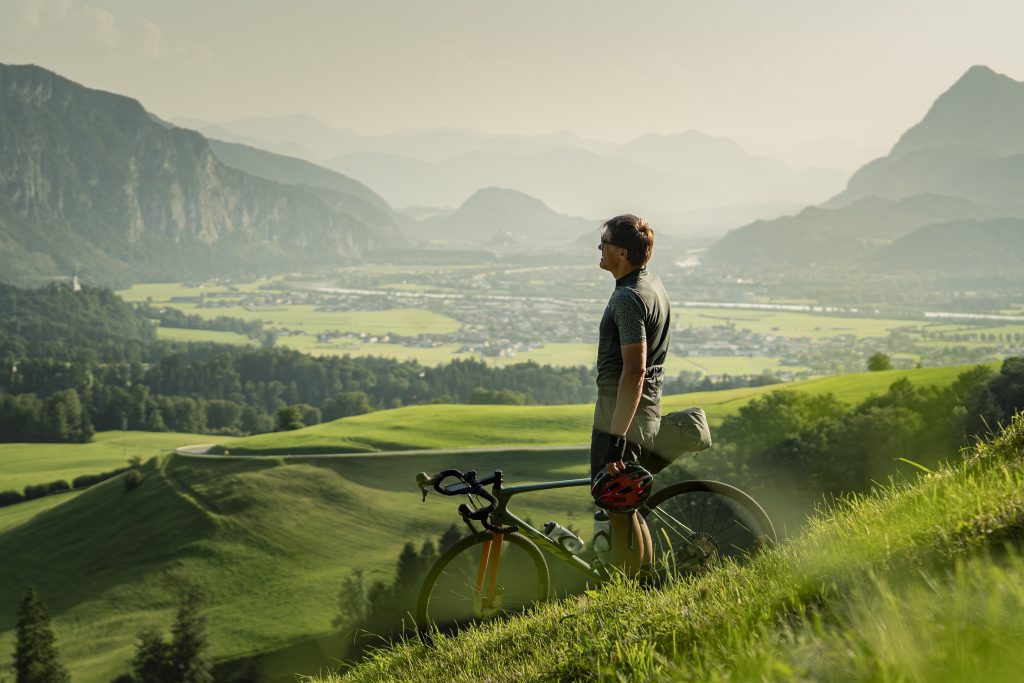Ein Mann in Radkleidung steht auf einer grünen Wiese neben seinem Gravelbike. Hinter ihm ist ein Fluss und Berge in der Landschaft zu sehen